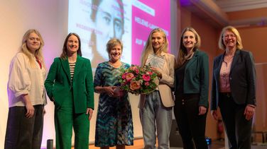 Das Bild zeigt sechs Damen, die vor einer Leinwand stehen, auf die das Logo des Helene-Lange-Preises projiziert wird. Sie lächeln in die Kamera. Die Preisträgerin Sandra Drolshagen hält einen Blumenstrauß und eine Urkunde in ihren Händen.