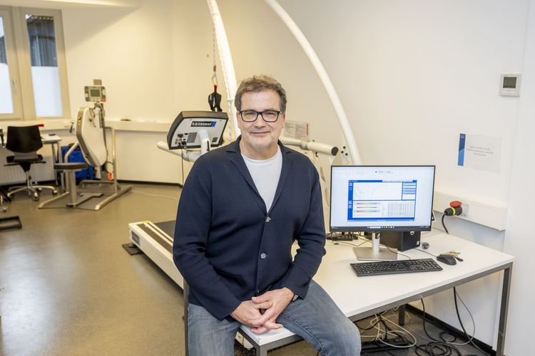 Das Foto zeigt Dirk Büsch. Er sitzt in einem Laborraum auf einem Tisch. Neben ihm steht ein Monitor und zeigt Forschungsdaten. Im Hintergrund kann man diverse Trainingsgeräte erkennen. Büsch lächelt in die Kamera.